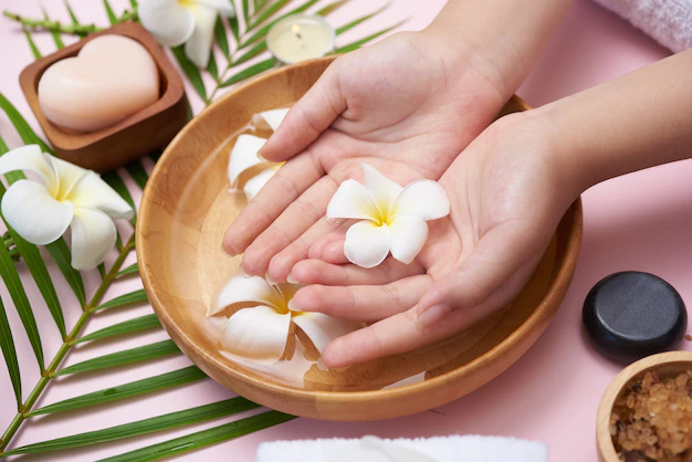 Woman soaking her hands in bowl of water and flowers, spa treatment and product for female feet and hand spa, massage pebble, perfumed flowers water and candles, relaxation. flat lay. top view.